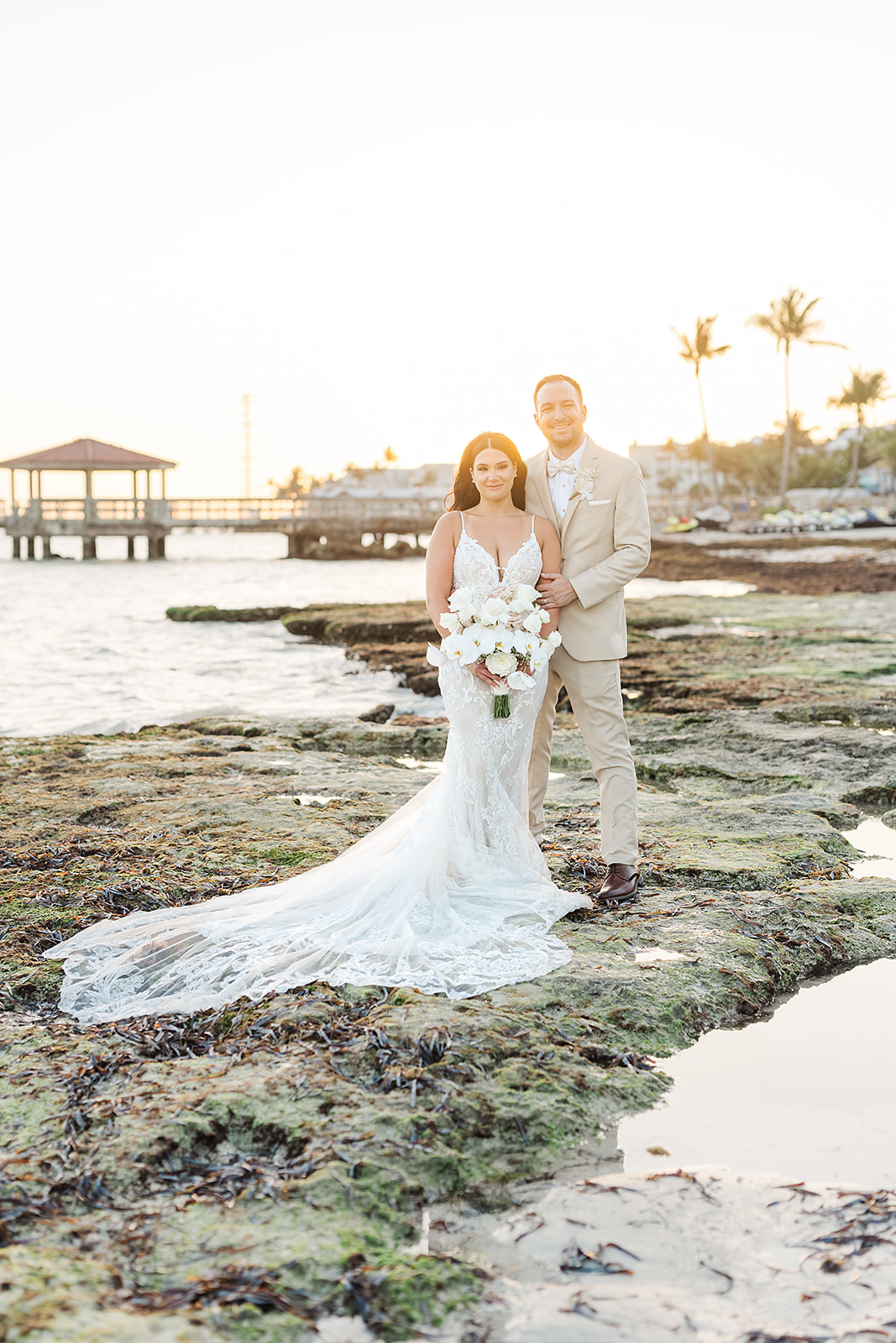 bride and groom pose for a wedding portrait on the beach next to tide pools with a cabana a pier behind them